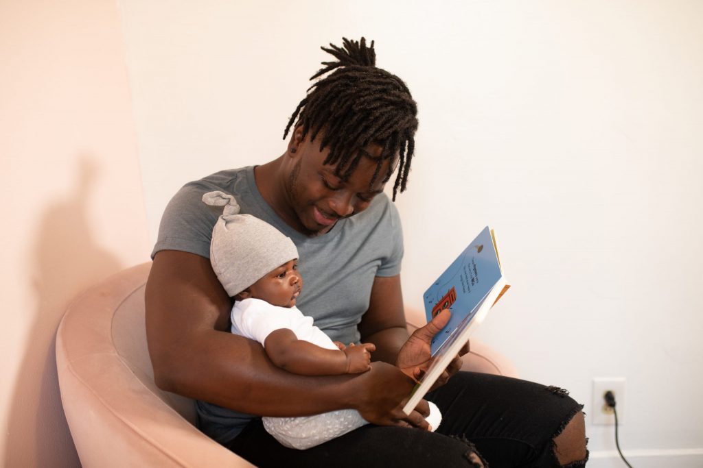 A Black father reads a book with their infant Black child.