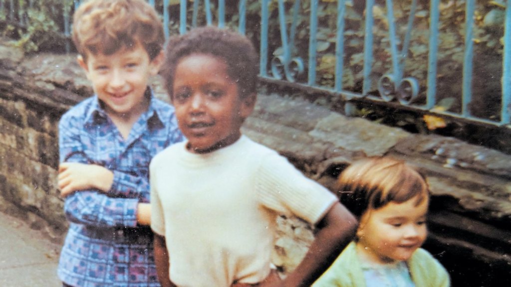 Three young children, two white, one Black stand infront of a wall with a blue fence on top. The young Black child stands in the middle, his hands on hips smiling at the camera.