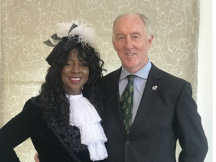 A Black woman wears a black and white feathered headpiece, and a ruffled white scarf over a black velvet jacket. This is the uniform of the High Sheriff, she stands next to her husband who is white and wears a dark blue suit and tie.