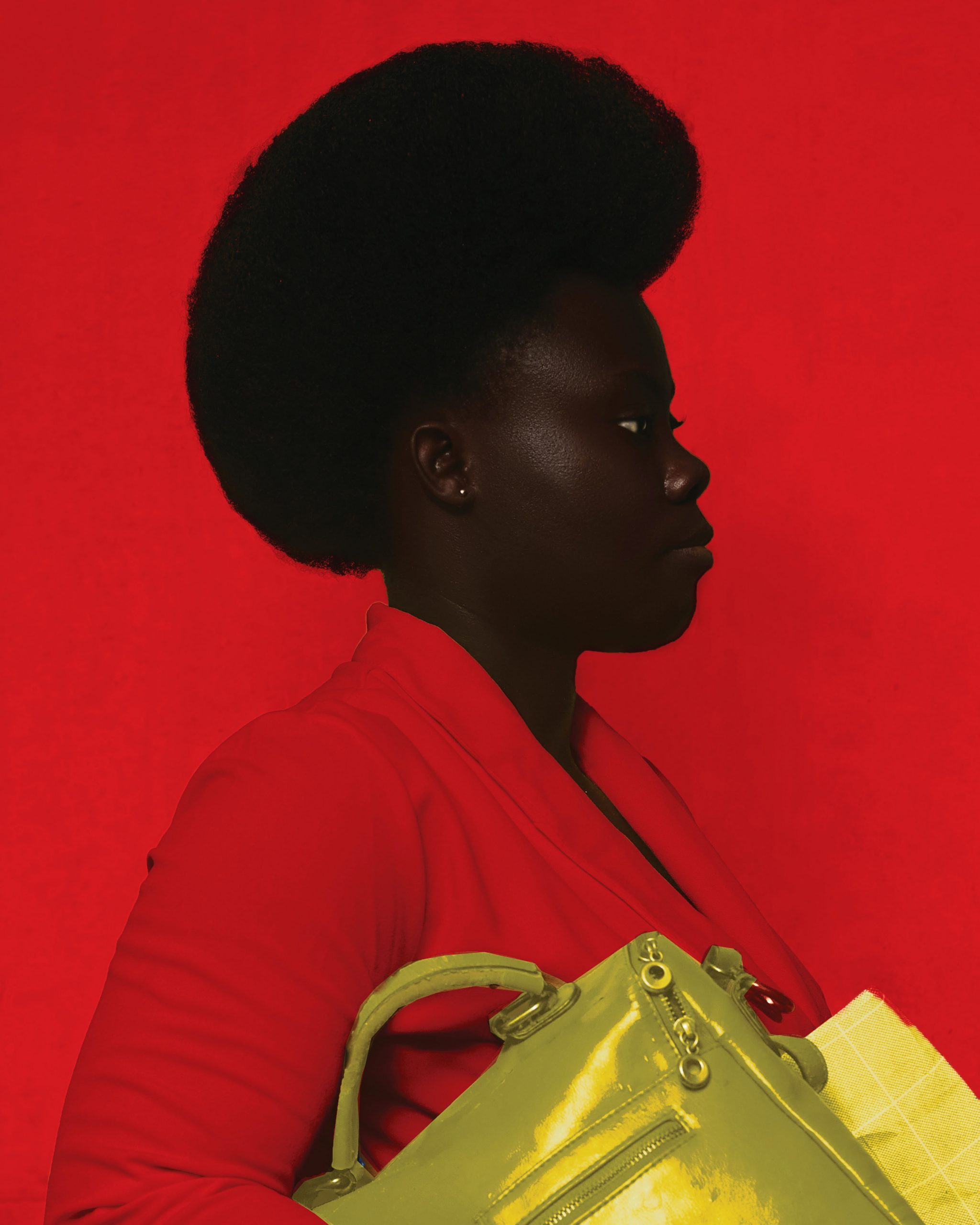 Profile of a dark skinned Black woman with shaped black afro hair wearing a red shirt and carrying a bright yellow handbag against a red background.