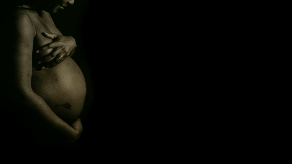 A naked pregnant Black woman can be seen from the waist up against a black backdrop. She is holds her arms over her breasts.