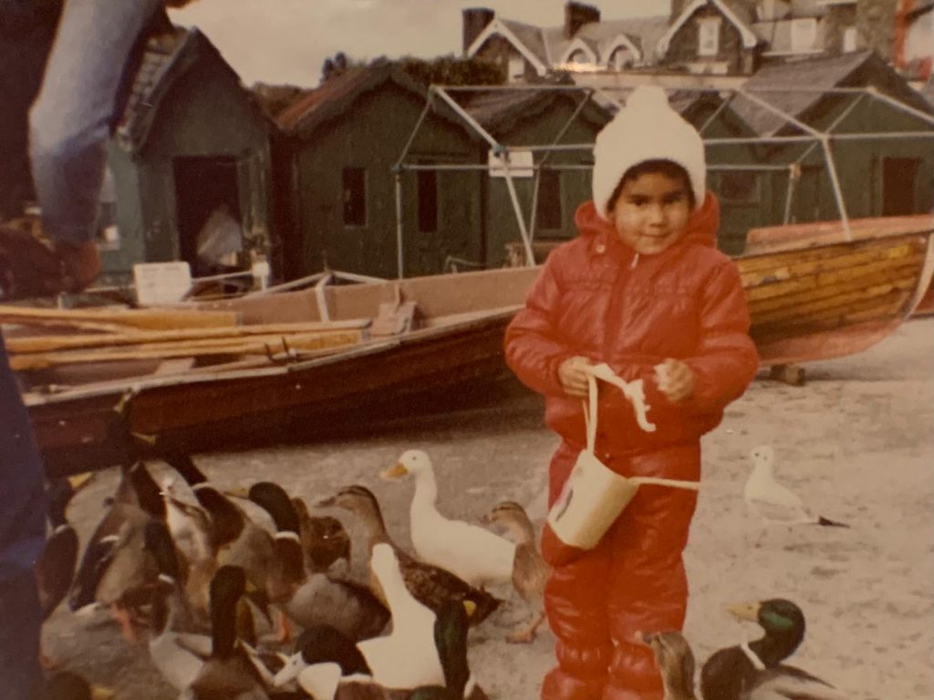 A young Black child in a red snowsuit and white bobble hat feeds geese.