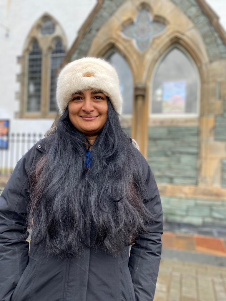 Zainab, a Brown woman with long dark hair wears a dark jacket and white hat.