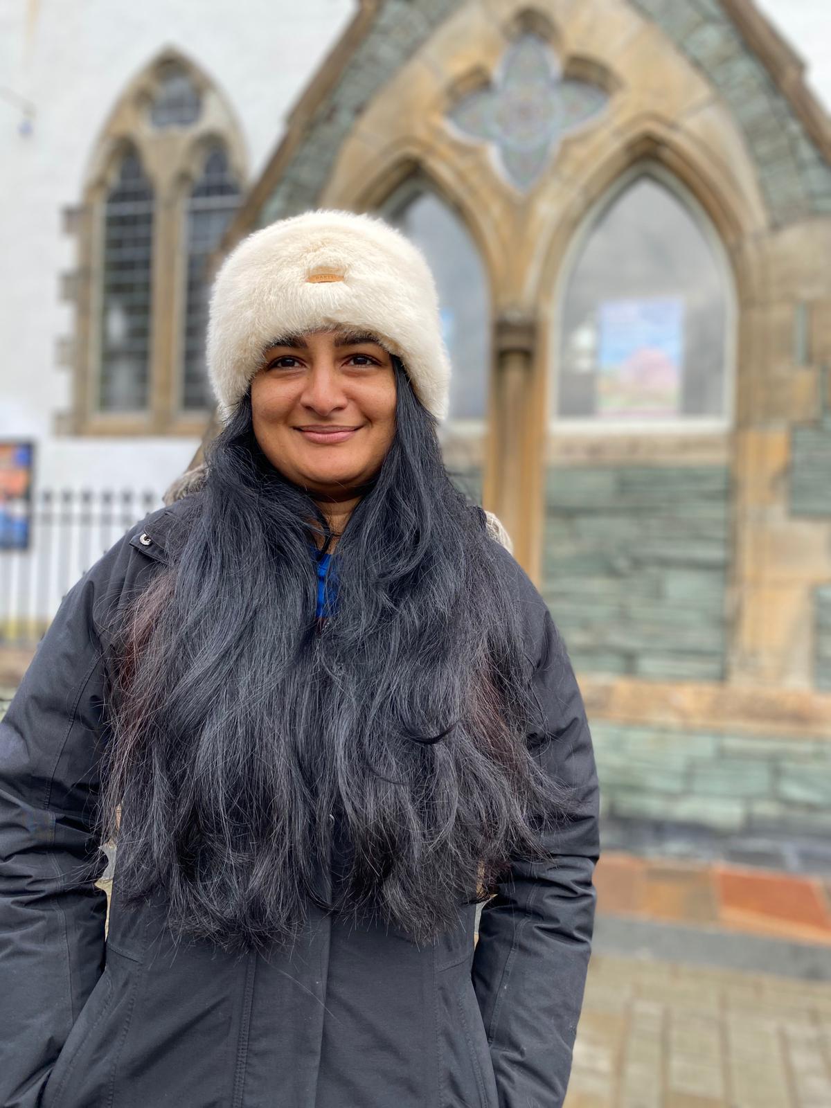 Zainab, a Brown woman with long dark hair wears a dark jacket and white hat.