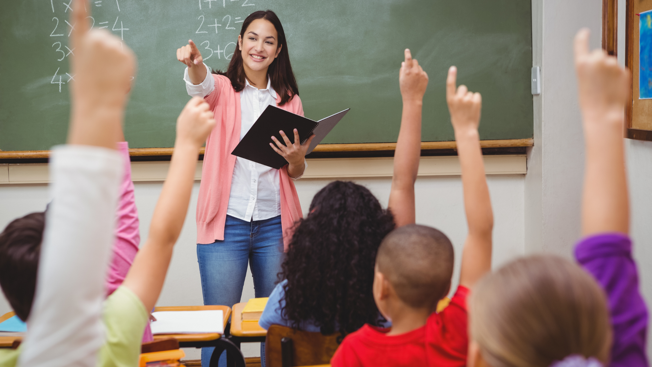 Image of a female teacher in a class of children with their hands up.