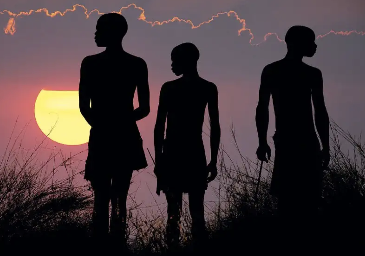 Three youth of African heritage stand in long grass with the sun set in the background.
