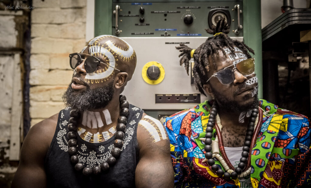 The lead singers of K.O.G & THE ZONGO BRIGADE: two Black men dressed in sunglasses, wooden beads, colourful clothing and white painted patterns on their skin.
