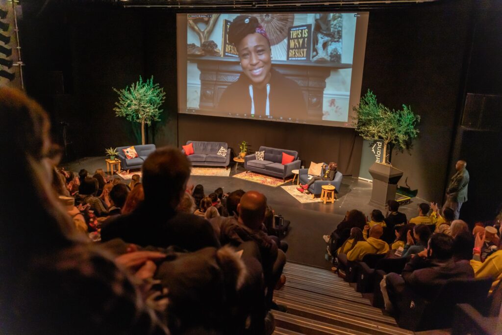 An audience at ARC Summit 2021 watch Dr Shola Mos-Shogbamimu on a large screen. Dr Shola is a Black woman wearing a black hoodie. She is smiling.