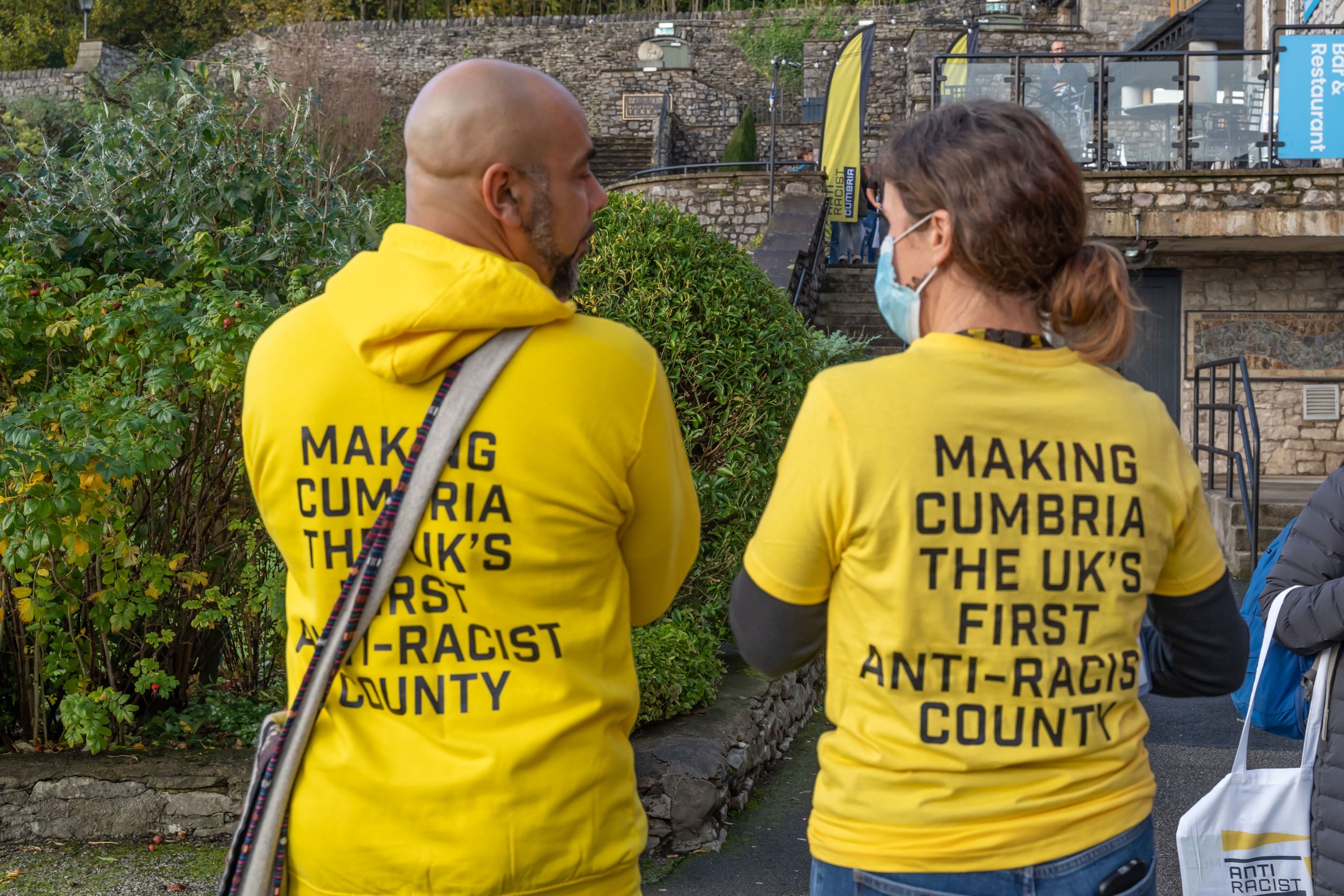 Two ARC volunteers; one Black man, one white woman wearing a mask, have their backs to the camera. They are wearing yellow ARC hoodies with black text: MAKING CUMBRIA THE UK'S FIRST ANTI-RACIST COUNTY