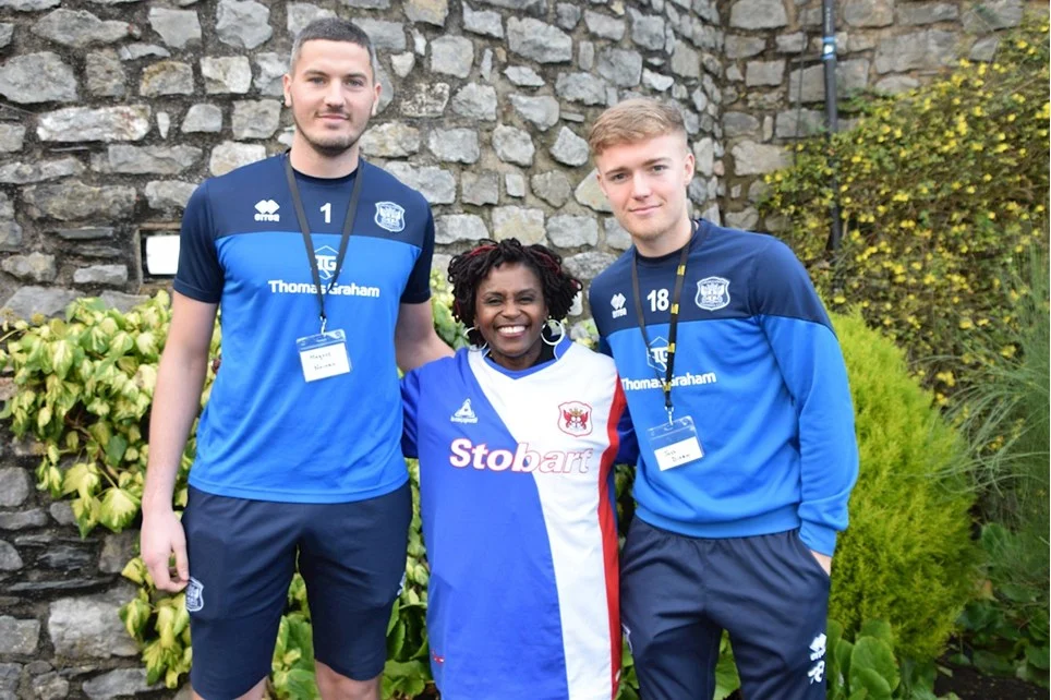 Janett, a short Black woman wears a Carlisle Utd FC shirt and stands between two white men in Carlisle Utd FC kit.