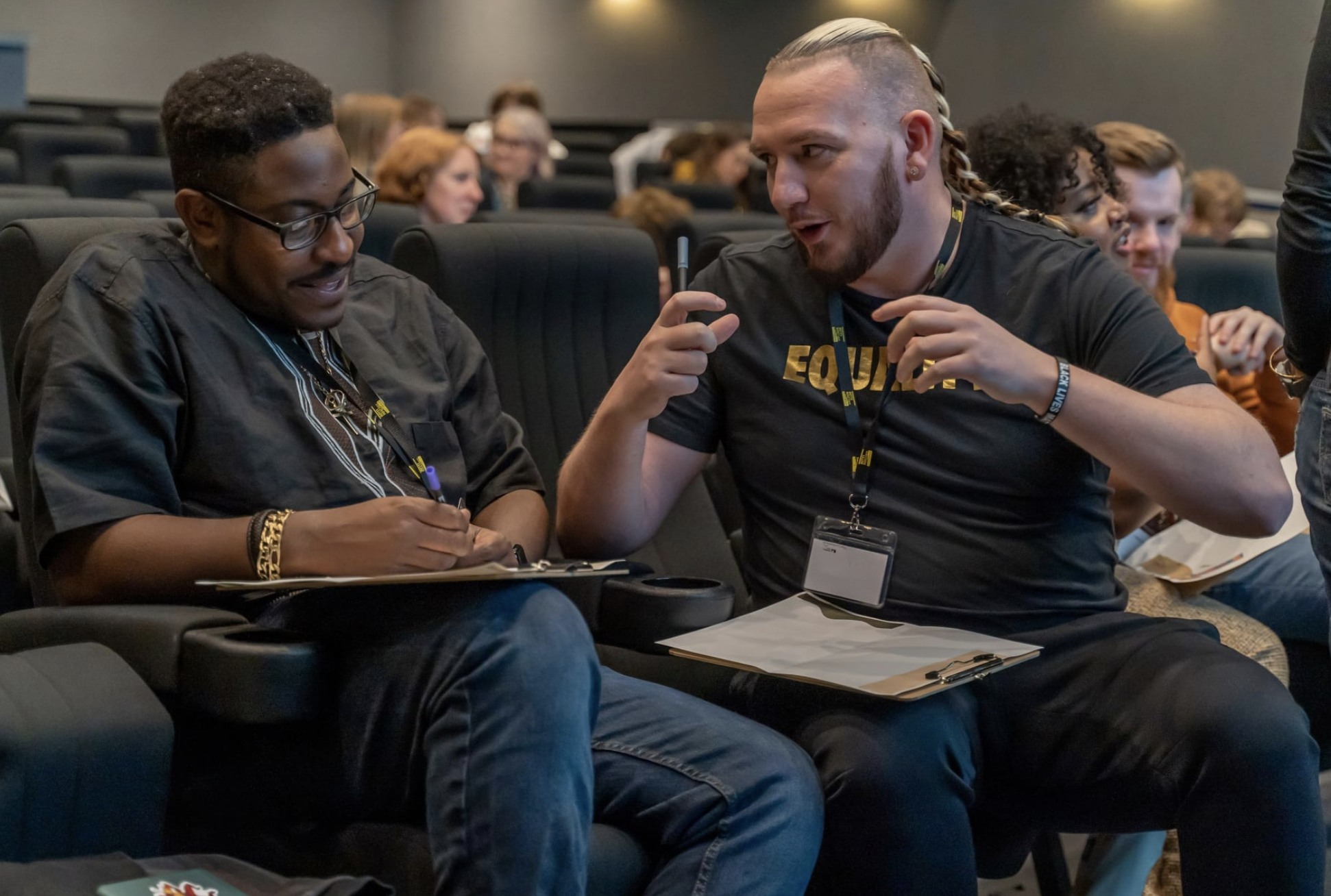 A black man and a white man are sitting in the audience having a conversation and smiling. They have clipboards and lanyards.