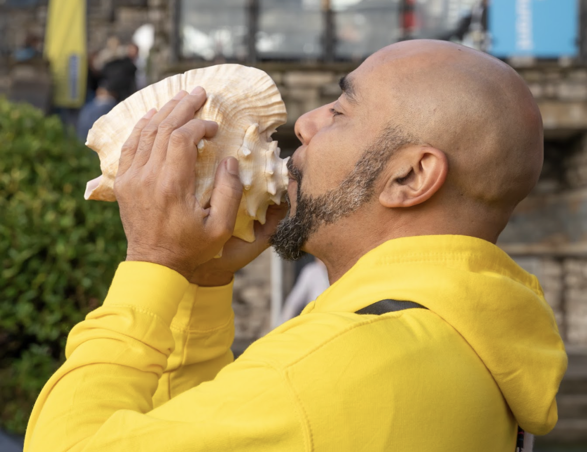 Middle-aged black man wearing a yellow hoodie blows a conch sea shell.