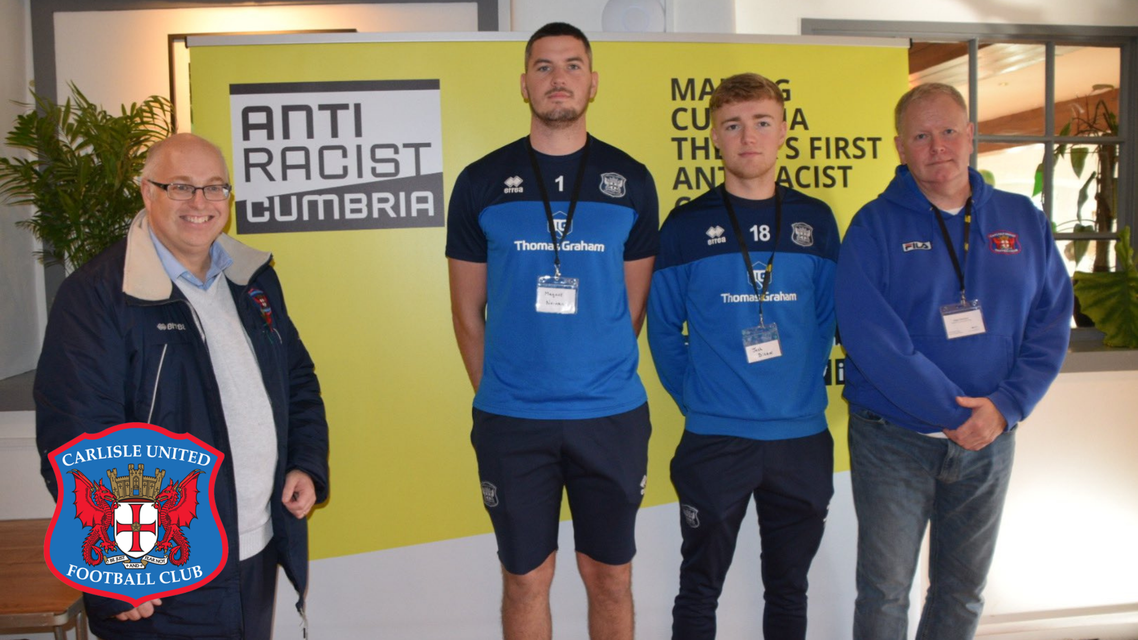 The CEO, two players and Media Manager of Carlisle Utd stand in a line in front of a large yellow Anti Racist Cumbria banner. The Carlisle Utd badge is overlayed to the bottom right of the image