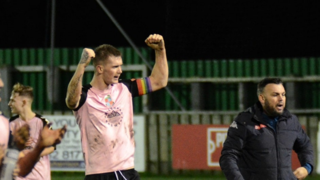 A white man in a pale pink sports shirt lifts his fists in celebration on a football pitch.