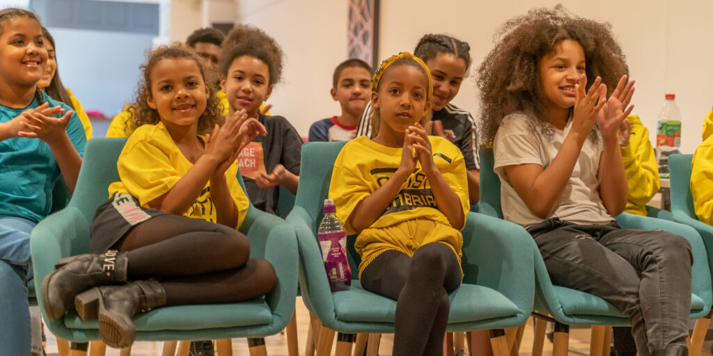A group of young black and brown children sitting down, smiling and clapping their hands.