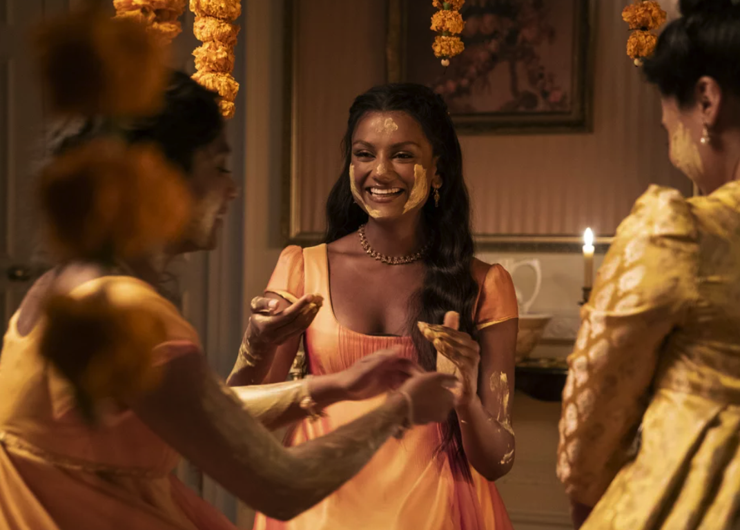 Three South Asian women in period dress smiling and clapping performing the Haldi ceremony; a South Asian wedding tradition, mixing turmeric with water and oil, spreading it on the body.