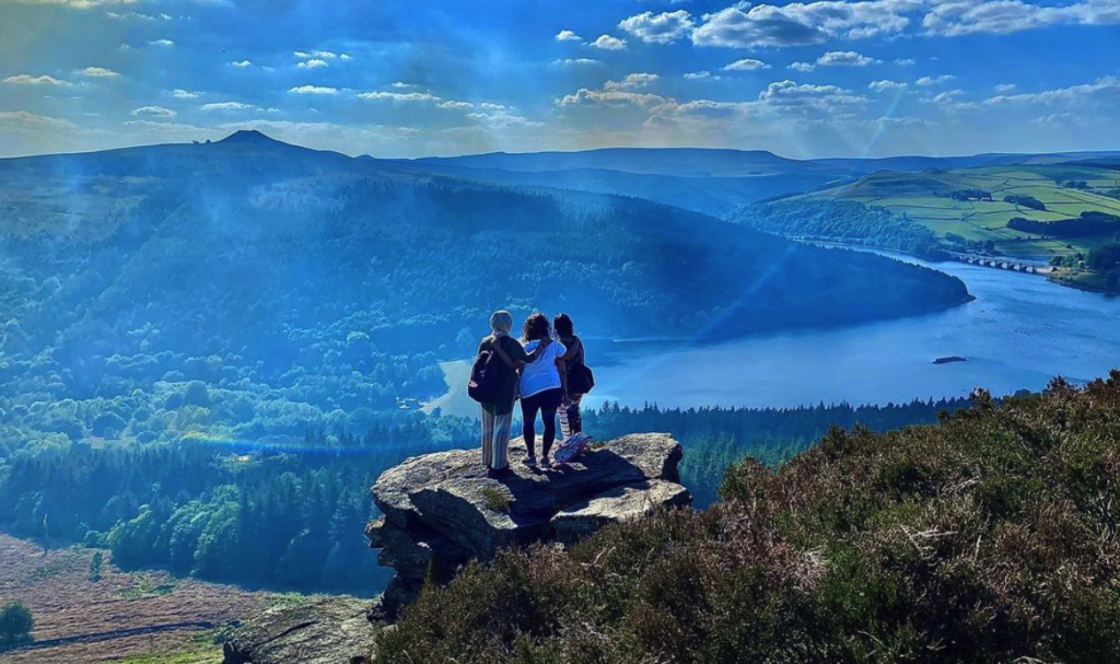 Three people stand on a rock on a sunny day overlooking a lake and fell. One person wears a headscarf, one has curly dark hair and one has straight dark hair.
