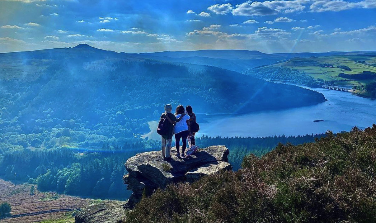 Three people stand on a rock on a sunny day overlooking a lake and fell. One person wears a headscarf, one has curly dark hair and one has straight dark hair.