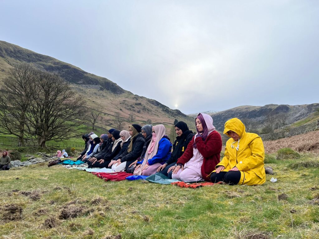 A group of women wearing headscarfs and jackets kneel down to pray on a hillside.