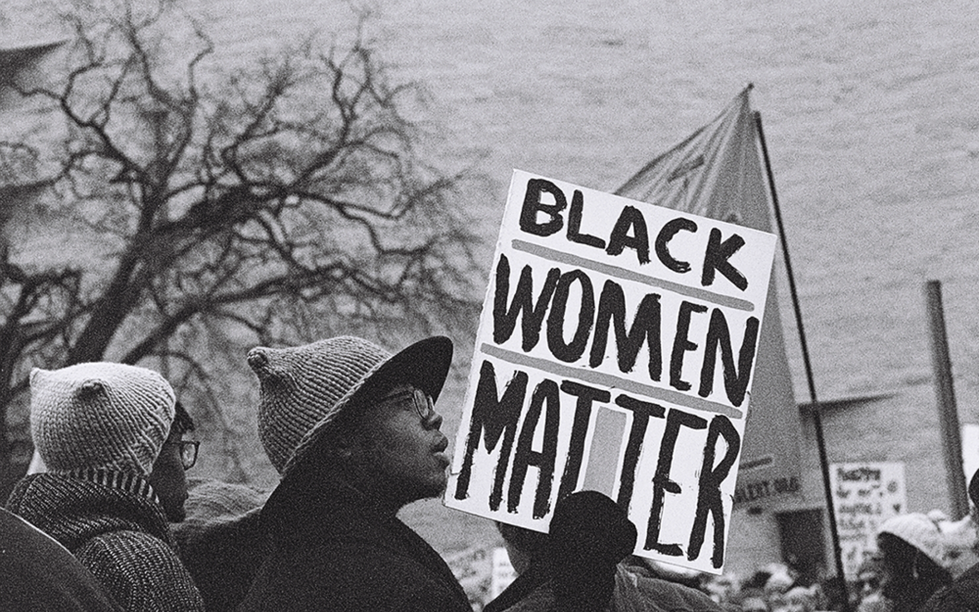 A black and white photo shows a section of a rally. A Black woman wearing a hat holds a sign that says BLACK WOMEN MATTER