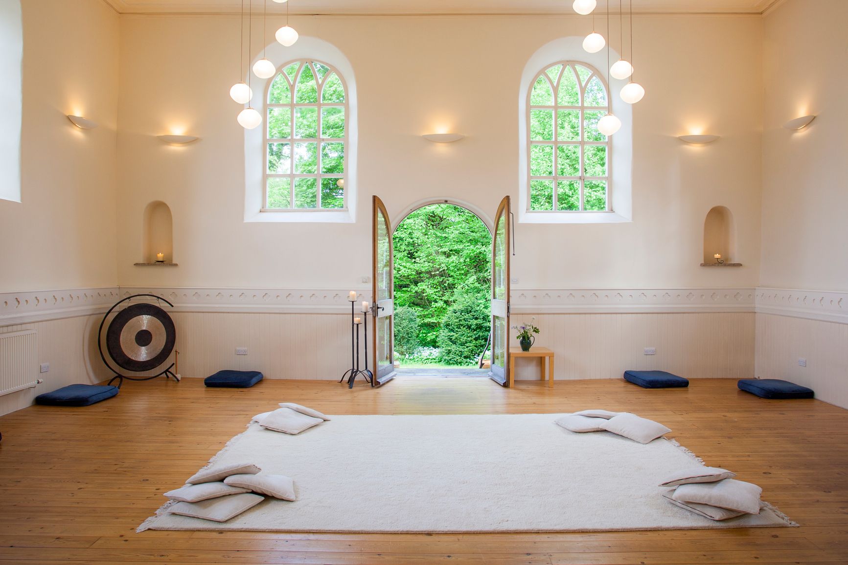 A large, well-lit room with two windows, white walls and wooden floor. There is a door open, a gong and a large rug on the floor with cushions in each corner.