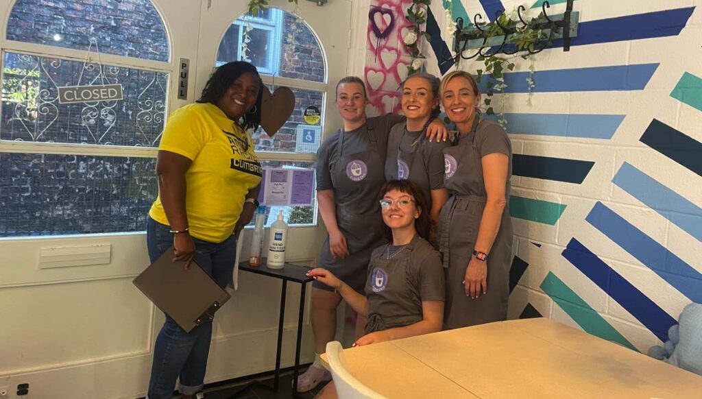 An black female Anti Racist Cumbria volunteer wears a yellow t-shirt and smiles next to four white café employees. There is an "ARCAngels" sticker on the café window.