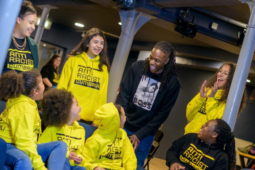 Group of black and brown children wearing yellow Anti-Racist Cumbria hoodies with Nigel Clarke, a black man. They are all smiling.