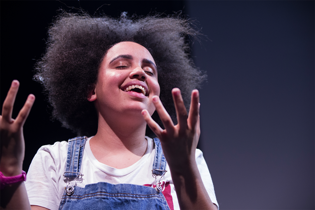 Cumbrian playwright Lekhani Chirwa is a mixed heritage woman with afro textured hair. She has her eyes closed and hands out. She is acting in her one-woman show "Can I Touch Your Hair?"