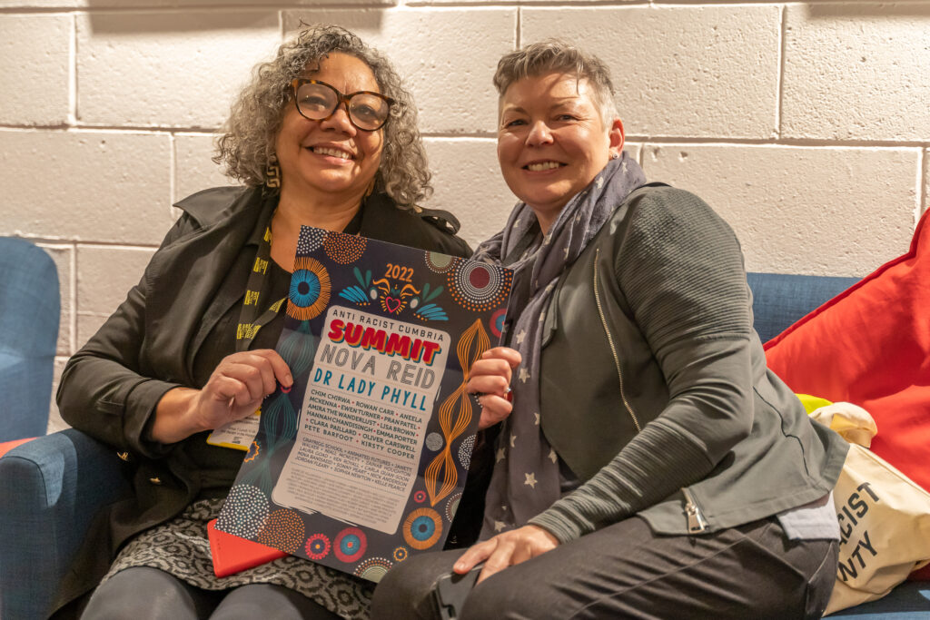 Two women sit on a sofa, they smile at the camera and hold up a poster of the event.