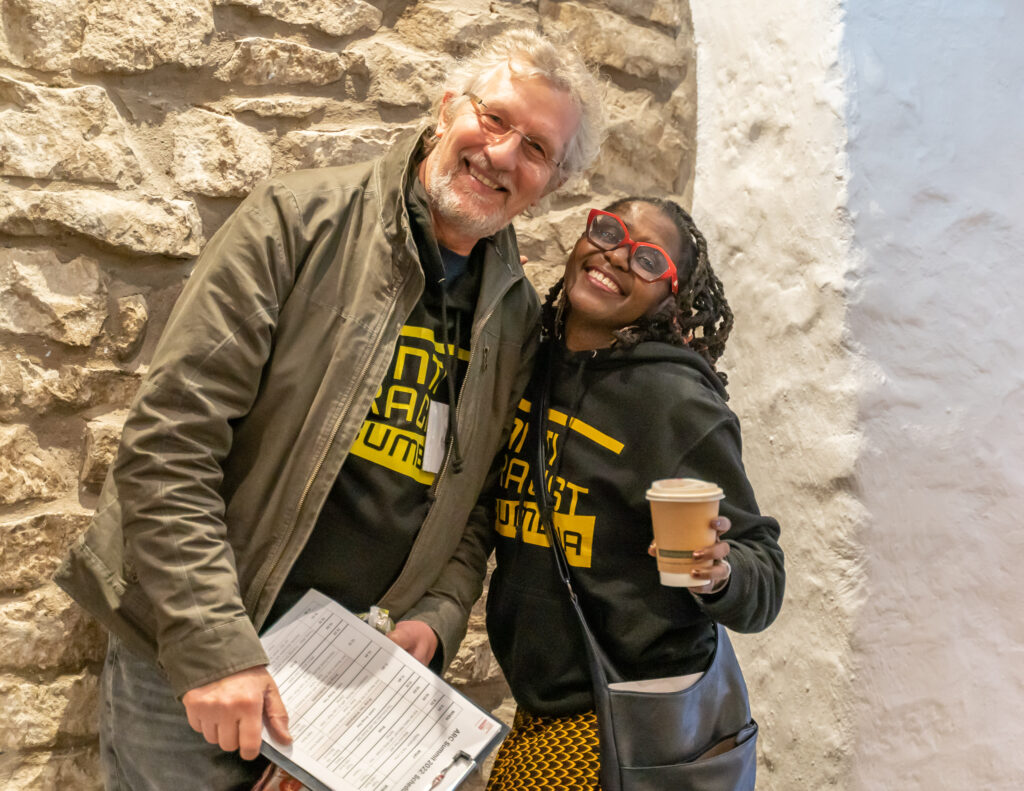 A white man and a Black woman stand smiling together. The man holds a clipboard, he has grey hair and wears a green jacket, the woman wears red glasses and holds a coffee. They both wear Anti Racist Cumbria hoodies.