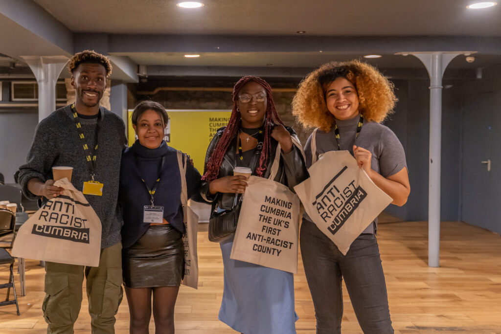 Four Black people smile at the camera. They hold totes bags which say 'Making Cumbria the UK's first anti-racist county.'
