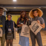 Four Black people smile at the camera. They hold totes bags which say 'Making Cumbria the UK's first anti-racist county.'