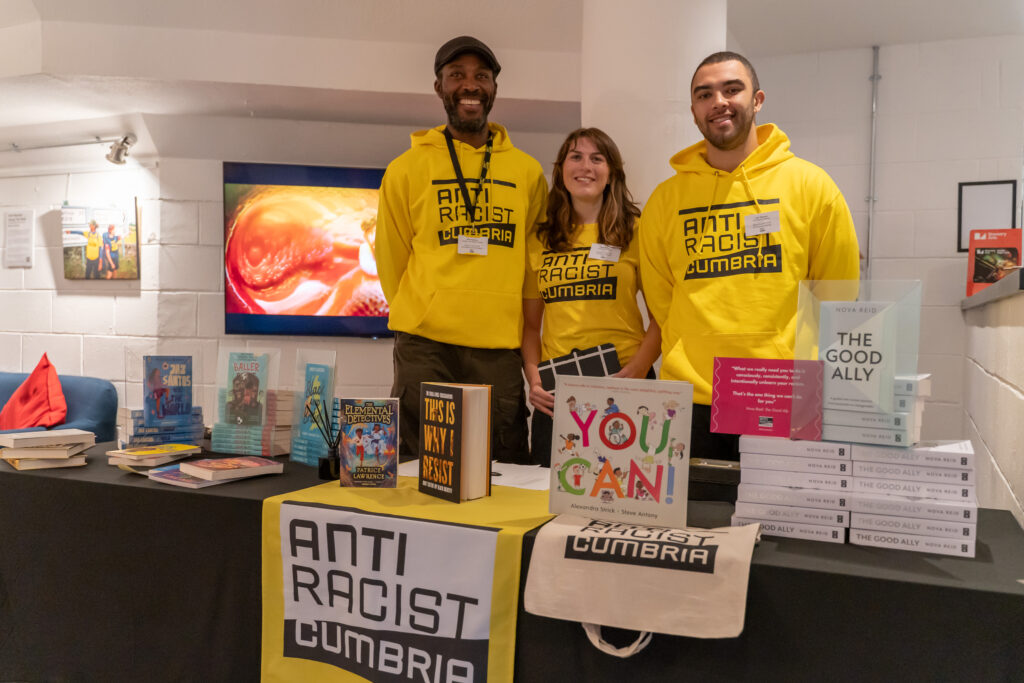 Three Anti Racist Cumbria volunteers stand behind the stand displaying books and merchandise for sale. The volunteers are two Black men and one white woman.