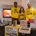 Three Anti Racist Cumbria volunteers stand behind the stand displaying books and merchandise for sale. The volunteers are two Black men and one white woman.