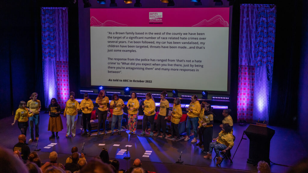 A row of people singing. In the background a story of someone's lived experience of racism is shared.