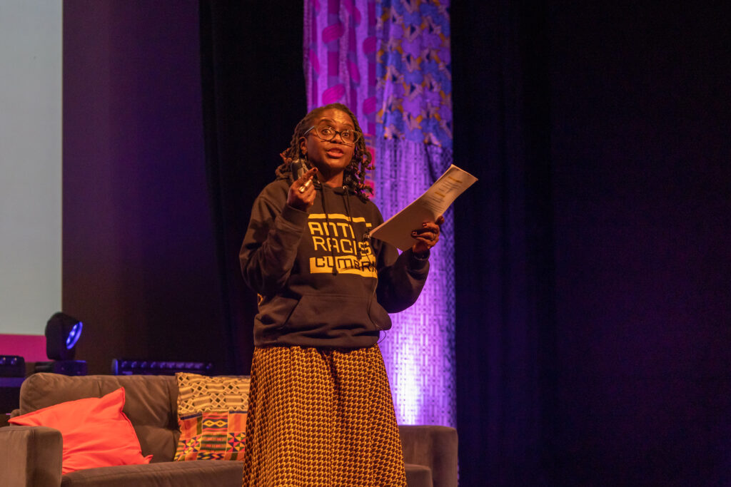 A Black woman talks on stage. She wears an Anti Racist Cumbria hoody and an African print skirt. Her locs are twisted up to a shoulder length.