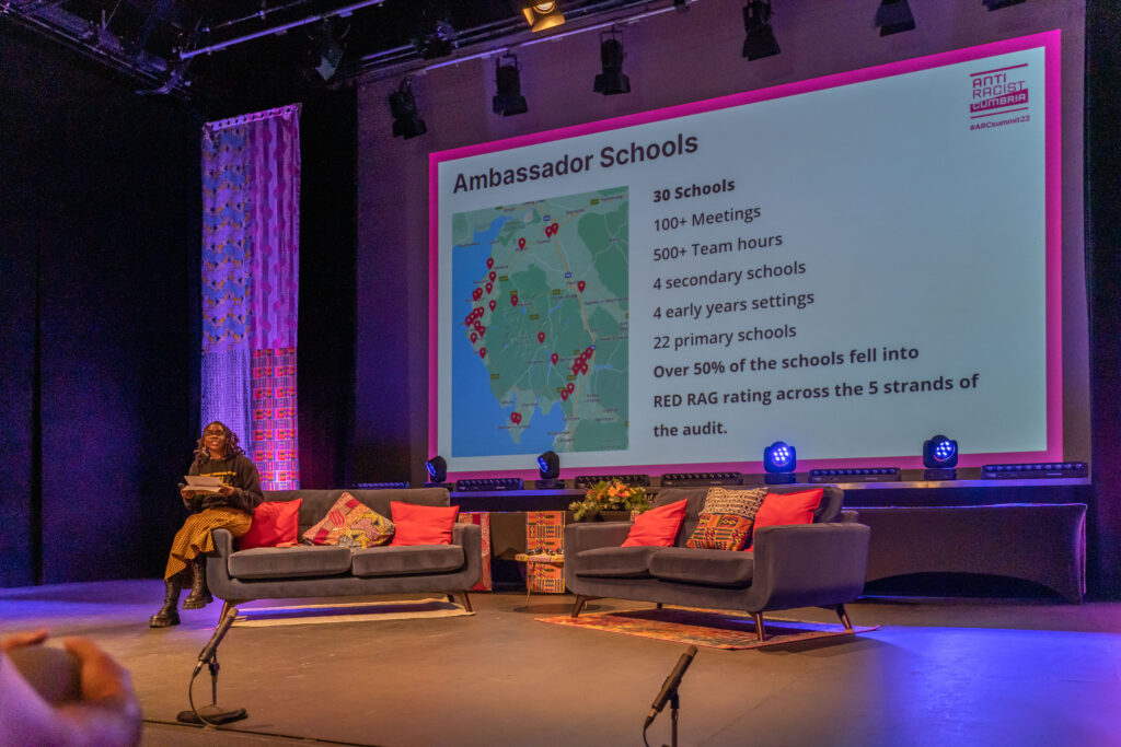 A Black woman talks on stage. She wears an Anti Racist Cumbria hoody and an African print skirt. Her locs are twisted up to a shoulder length.