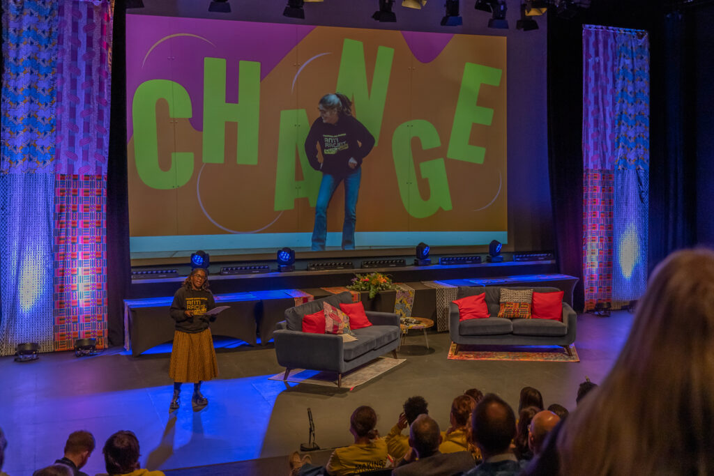 A Black woman talks on stage. She wears an Anti Racist Cumbria hoody and an African print skirt. Her locs are twisted up to a shoulder length. In the background a colourful photo shows a white woman wearing the same hoody in front of a vibrant wall which has the word CHANGE on it.