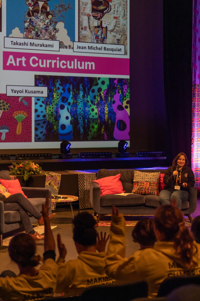A white woman sits on a sofa, she talks into a microphone. Behind her a screen shows a variety of artworks by artists who aren't white.