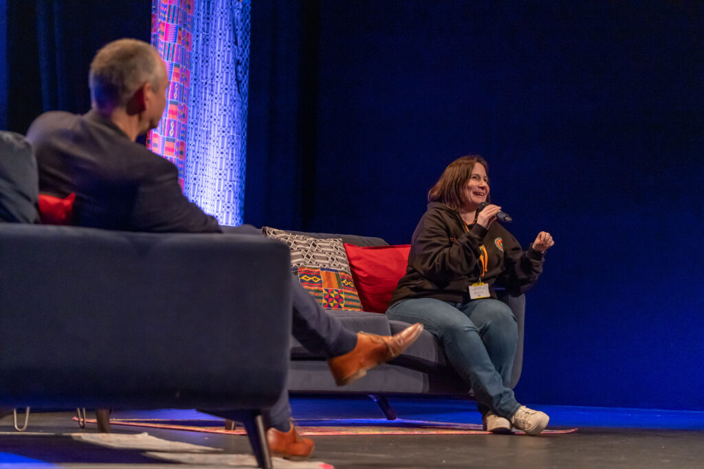 A white woman smiles as she talks into a microphone, the back of a white man can be seen in the foreground.