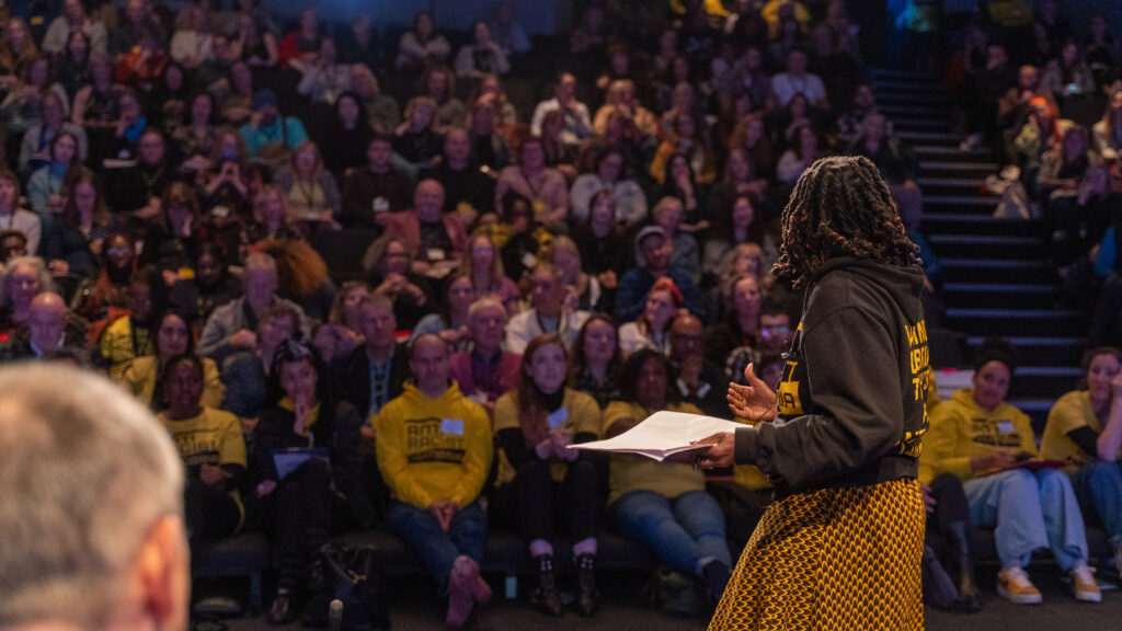 A full auditorium can be seen over the shoulder of a Black woman.
