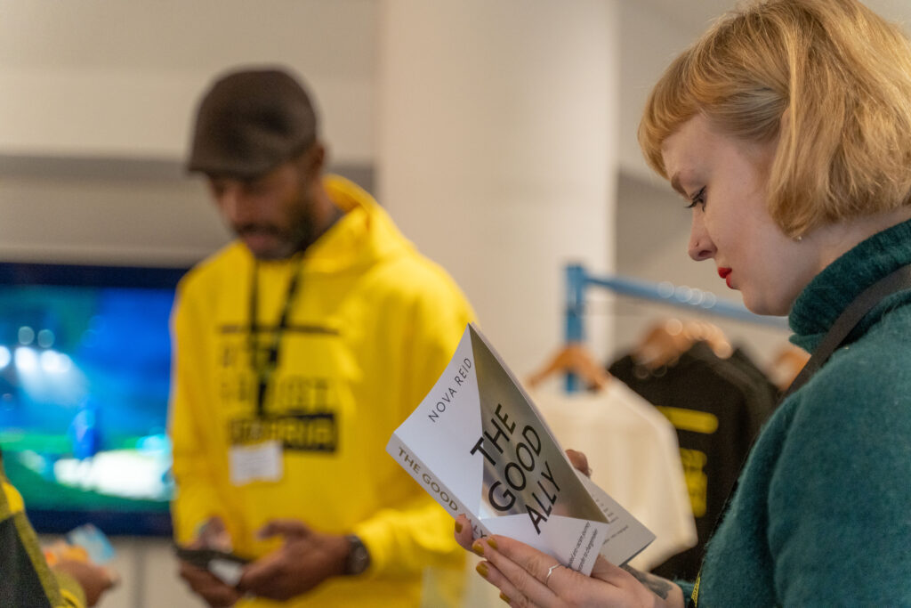 A white woman is reading a book called The Good Ally by Nova Reid. In the background a Black man wearing an Anti Racist Cumbria hoody is selling someone something at a stand.