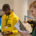 A white woman is reading a book called The Good Ally by Nova Reid. In the background a Black man wearing an Anti Racist Cumbria hoody is selling someone something at a stand.