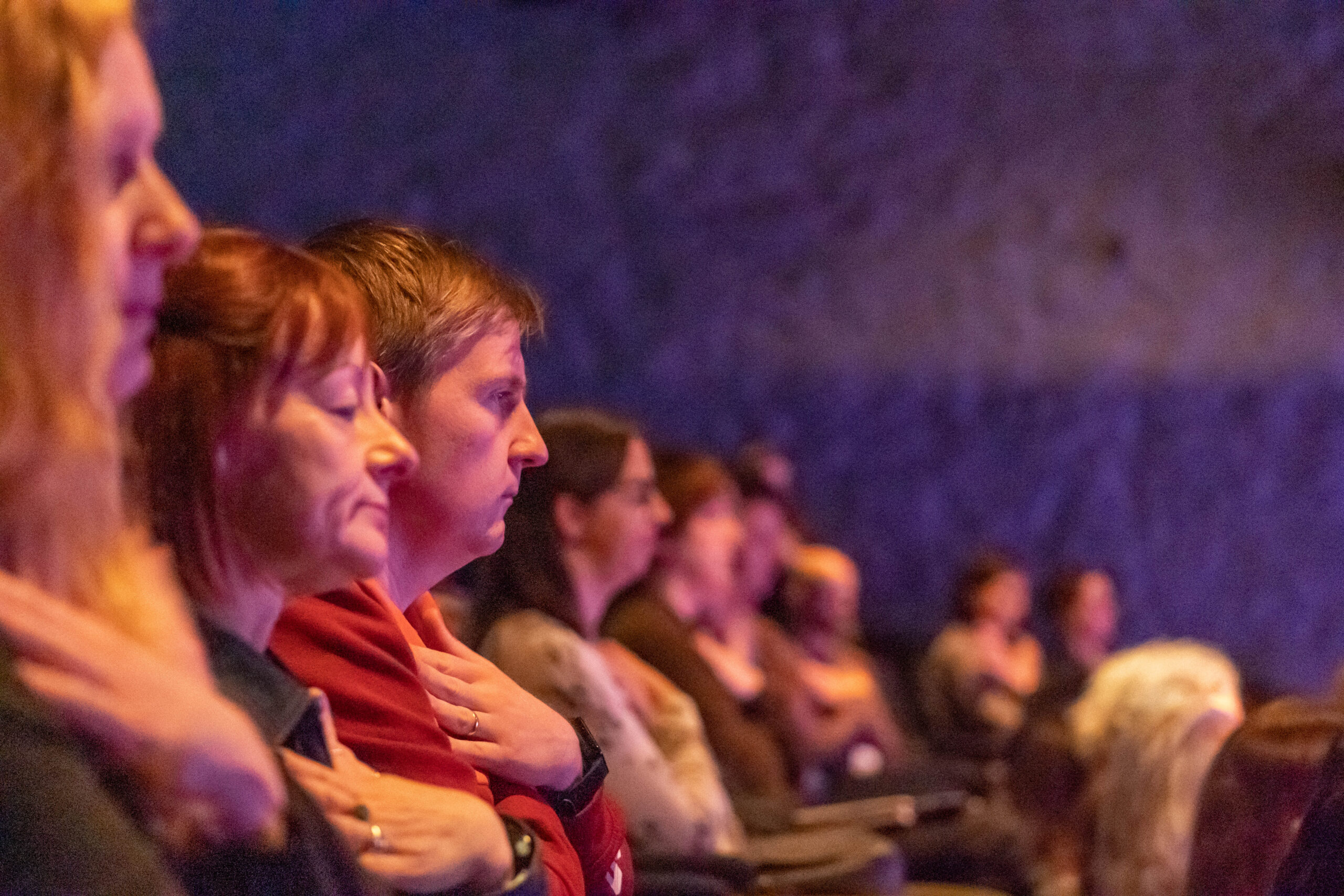 A row of people in an audience hold their hand on their heart, their eyes are closed.
