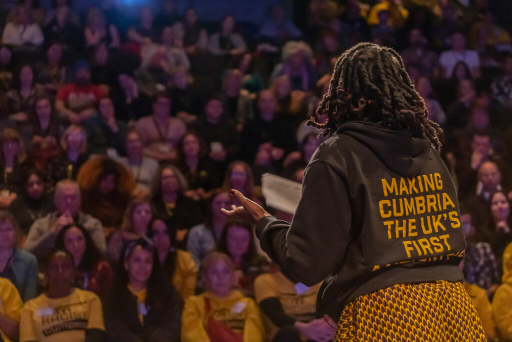 A full auditorium can be seen over the shoulder of a Black woman.