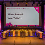 A large U-shaped table has black tablecloths and African print fabric banners hanging over this. There is a floral arrangement in the centre. A screen behind reads 'Who's Around Your Table?'