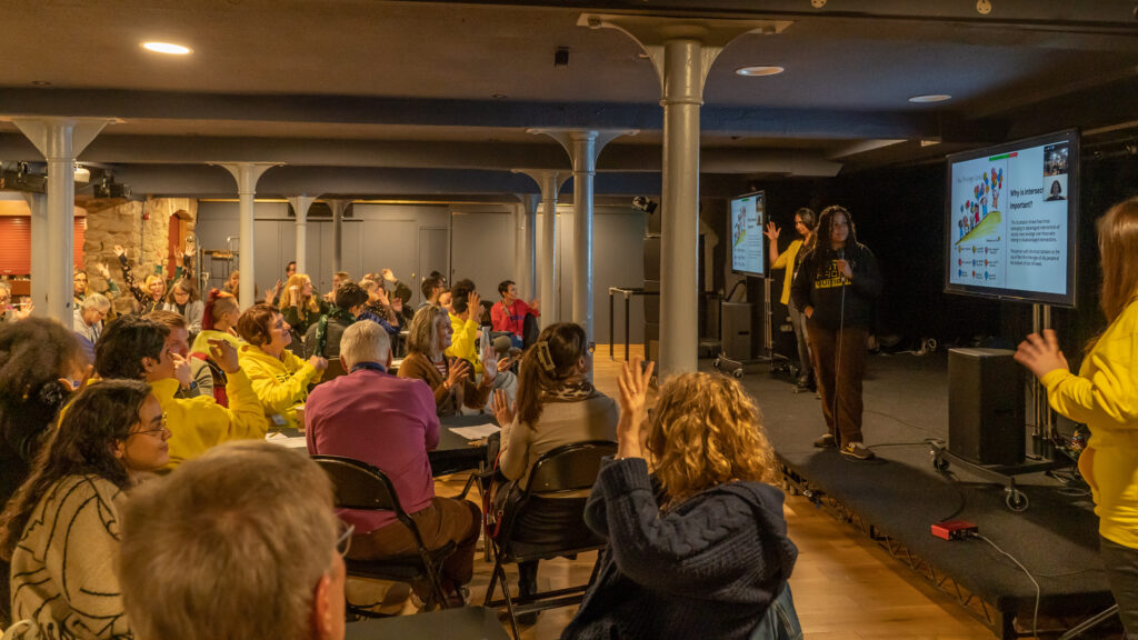 A room of people sitting around tables looking at the stage where a Black woman talks. They raise their hands in appreciation.
