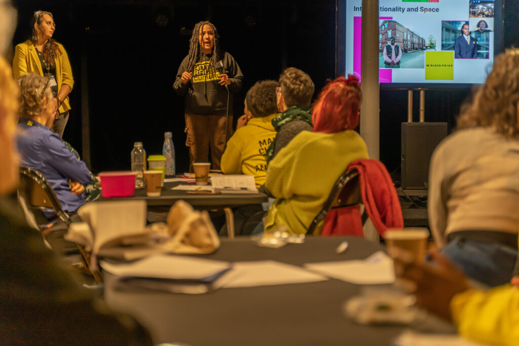 A Black woman wearing their hair in long braids gives a talk. In the foreground people look a tthe stage and listen.