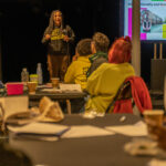 A Black woman wearing their hair in long braids gives a talk. In the foreground people look a tthe stage and listen.