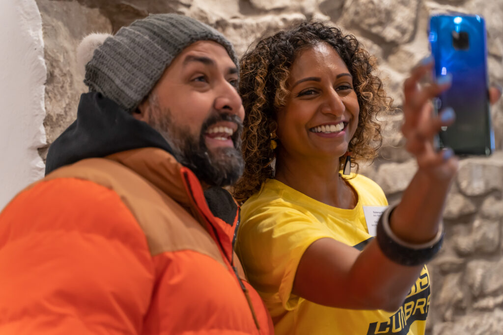 Two people with South Asian heritage take a selfie. One is a man with a beard wearing a bobble hat and orange puffer jacket, the other is a woman with a yellow t-shirt and curly hair.